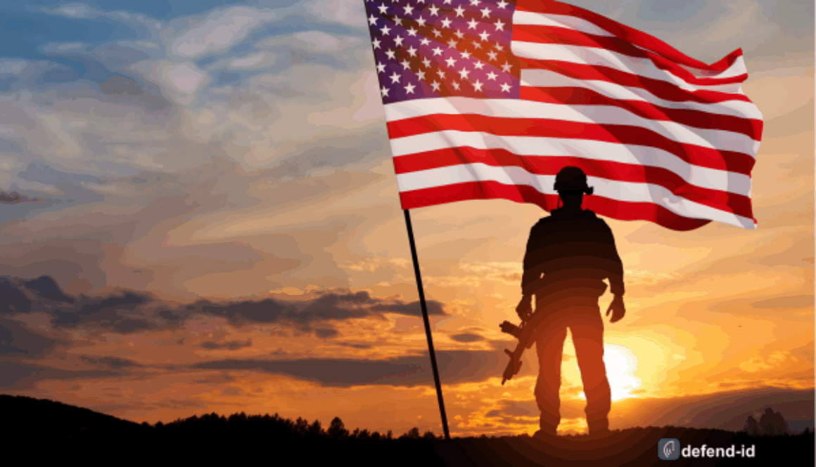 Silhouette of a soldier standing on a hill with the American flag waiving. The backdrop is an orange glowing sunset